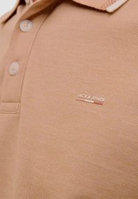 Bežs polo krekls ar tekstūrētu audumu, divām pogām pie apkakles un mazu metāla zīmogu ar uzrakstu "JACK & JONES."