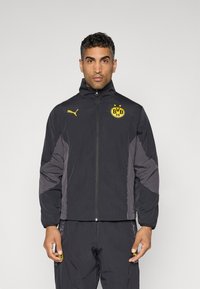 Schwarze leichte Jacke mit hohem Kragen, Reißverschluss und gelbem Puma-Logo. Mit grauen Seitenpanelen und BVB-Logo auf der Brust.