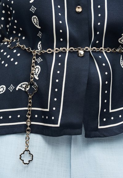 Μπλε σκούρο πουκάμισο με λευκό μοτίβο με σχεδιασμό παϊέτας και διακοσμητική τελεία στα σύνορα, ασημένιο κουμπί και ασημένια ζώνη με αλυσίδα με κράμα τετράφυλλων μοτίβων πάνω σε ανοιχτό μπλε παντελόνι.