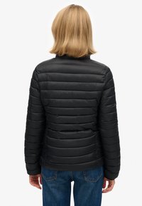 Czarna pikowana kurtka z poziomym wzorem, wysokim kołnierzem i gładką fakturą, zestawiona z niebieskimi dżinsami. Widok od tyłu.