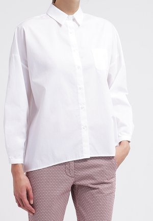 Person iført en løs hvid skjorte med knapper og brystlomme samt mønstrede, tætsiddende bukser med hånden i lommen.