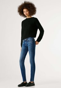 Mustast kootud ümar kaelusega kampsun, siniste kitsaste teksadega. Modell kannab musta tekstuuriga jalanõusid, seisab ühevärvilise tausta ees.