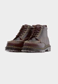 Ζευγάρι καφέ δερμάτινων μποτών αστραγάλου με χοντρές σόλες, αντίθετη ραφή και κορδόνια μπροστά σε γκρι φόντο.