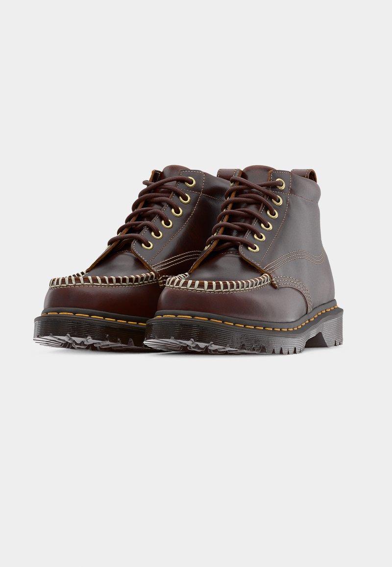 Paartje bruine leren enkellaarzen met dikke zolen, contrasterend stiksel en veters aan de voorkant op een grijze achtergrond.