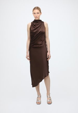 Femme portant une robe en satin marron foncé sans manches avec un ourlet asymétrique et des sandales à talons hauts argentées à brides, debout devant un fond uni.