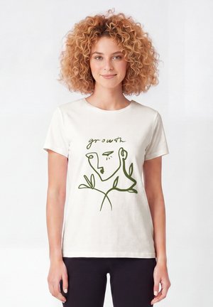 Vrouw met krullend haar die een wit t-shirt draagt met een groen abstract gezicht en bladontwerp en het woord "growth" erboven.