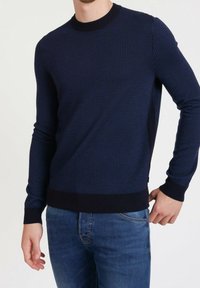 Pull en maille bleu marine avec un motif texturé, col rond, et poignets et ourlet côtelés. Porté avec un jean bleu et un logo discret au niveau de l'ourlet.