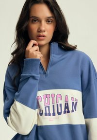 Niebieska bluza z zamkiem błyskawicznym z białymi i niebieskimi sekcjami w stylu color-block, z napisem "MICHIGAN" w różowym. Szczegóły w formie prążkowanych mankietów i kołnierza.