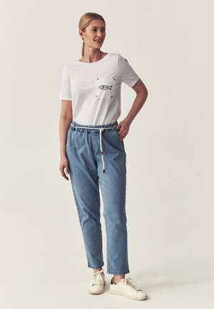 Frau stehend, trägt ein weißes T-Shirt mit einem Fischmotiv auf der Tasche, blaue Jeans mit Kordelzug und weiße Turnschuhe vor einem einfarbigen Hintergrund.