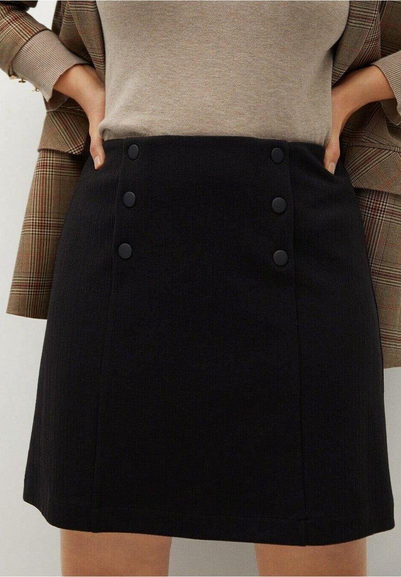 Femme portant une jupe noire avec six boutons mats et un haut beige, les mains posées sur les hanches, avec un blazer à carreaux visible derrière.