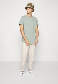 T-shirt vert clair, pantalon beige, chapeau seau camouflage et baskets blanches avec accents verts. Coupe décontractée et texture en coton douce.
