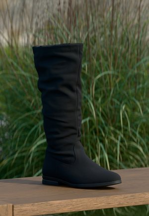 Tall black slouchy flat boot standing on a wooden bench with tall grass in the blurred background.