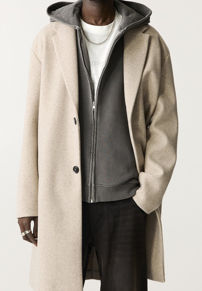 Homme portant un manteau en laine beige par-dessus un sweat à capuche zippé gris foncé et une chemise blanche, avec un collier en chaîne argentée et des bagues.