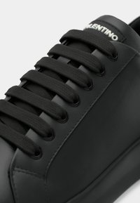 Baskets en cuir noir avec lacets plats noirs, œillets métalliques et un logo blanc sur la languette. Texture lisse avec un design à bout arrondi.