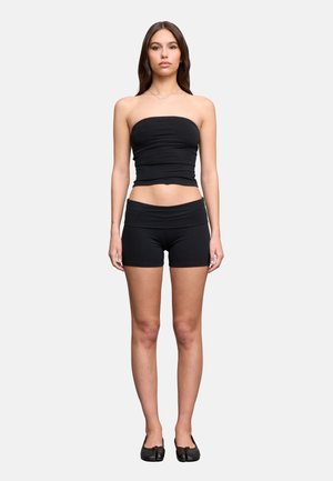 Mujer joven de pie, con un top negro sin tirantes, pantalones cortos negros ceñidos y zapatos planos negros contra un fondo liso y claro.