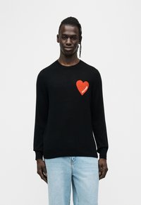 Pull en tricot noir à manches longues, avec un emblème en forme de cœur rouge sur la poitrine gauche portant le mot "FUTURE" en blanc.