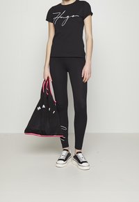 Schwarzes T-Shirt mit weißem Logo, schwarze Leggings und schwarze Tasche mit roten Akzenten. Fußbekleidung: schwarz-weiße Sneakers mit dicker Sohle.
