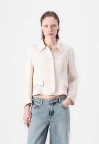 Kratka svetlo bež jakna s ostrim ovratnikom, pet velikih gumbov in sprednjimi žepi, v kombinaciji s svetlo modrimi kavbojkami. Gladka tekstura.