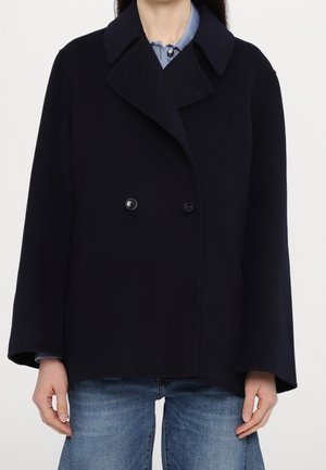 Femme portant un manteau croisé bleu marine foncé sur une chemise bleu clair et un jean bleu, debout devant un fond uni.