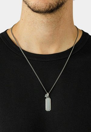 Collana d'argento con un ciondolo rettangolare caratterizzato da un bordo testurizzato, sospeso a una catena fine, indossata sopra una camicia nera.