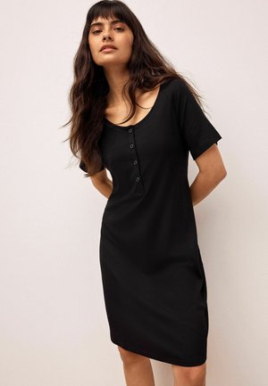 Woman with long dark hair wearing a black short-sleeve buttoned knit dress, standing with hands behind back against a plain background.