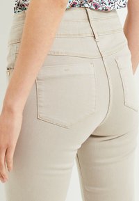 Gros plan sur une personne portant un pantalon cintré beige avec des poches arrière et un haut à motifs colorés rentré.