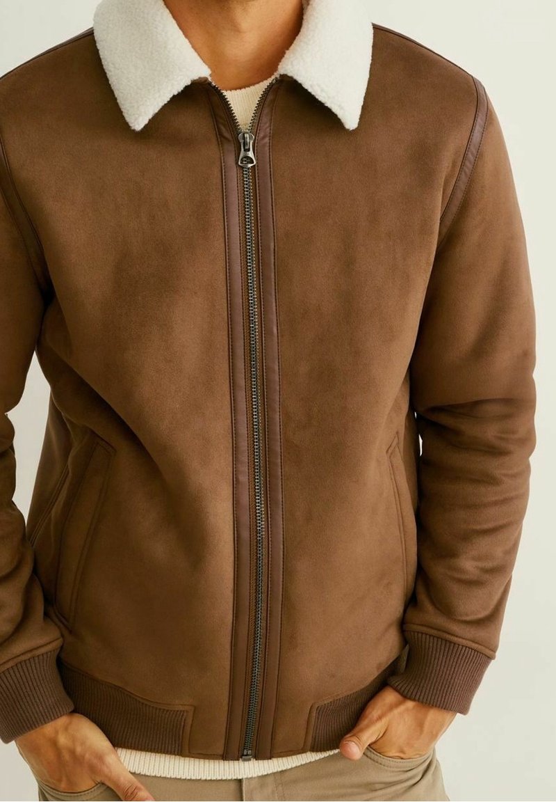 Uomo che indossa una giacca bomber in suede marrone con un colletto in pelliccia sintetica bianco, zip frontale e polsini e orlo a coste.