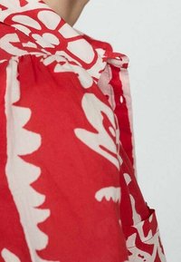 Camicia con motivo rosso e bianco caratterizzata da disegni floreali, con colletto e tessuto arricciato alle spalle. Materiale morbido e leggero.