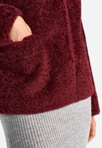 Une main émerge d'une poche d'une veste en peluche bourgogne, associée à une jupe grise côtelée, mettant l'accent sur le contraste de textures et de couleurs.