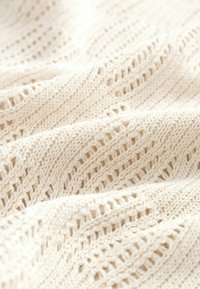 Beige strikket stof med et hulmønster. Blød tekstur med skiftende rækker af større og mindre huller i et bølgende design.