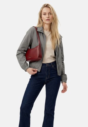 Lyshåret kvinde iført grå lynlåstrøje, beige top, mørkeblå jeans og bærende på en lille mørkerød skuldertaske.