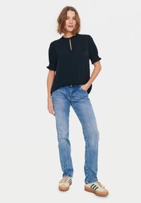Blusa blu navy con scollatura a chiave e maniche a sbuffo, abbinata a jeans azzurri chiari e sneakers beige con dettagli verdi.