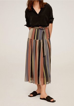 Vrouw draagt een zwart overhemd met knopen, een midi-rok met multicolor verticale strepen en een strikceintuur, en zwarte slippers, staat met handen achter de rug.