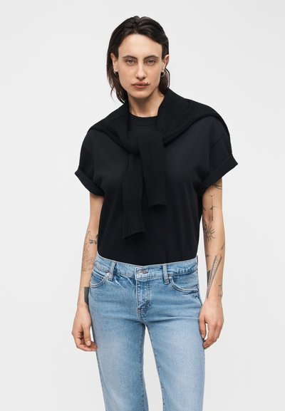 Jeune personne portant un t-shirt noir à manches retroussées avec un pull noir posé sur les épaules et un jean bleu clair, montrant des tatouages sur le bras.