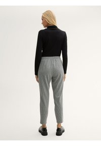 Schwarzes Langarmshirt und graue karierte Hose mit elastischem Bund. Die Schuhe haben dicke, schwarze Sohlen. Glatte und strukturierte Materialien.