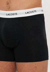 Fekete pamut boxer alsónadrág fehér elastikus derékkal, melyen ismétlődő fekete "LACOSTE" logó található. Simább textúra és testhezálló fazon.