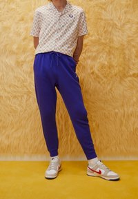 Kortärmad skjorta med geometriskt mönster i krämfärg och röd, kombinerad med blåa sweatpants och neutrala sneakers med röda och gröna detaljer.