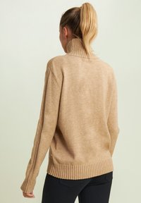 Pull en tricot beige avec un col montant, ourlets et poignets côtelés, présentant des rayures verticales le long des manches et une texture douce.