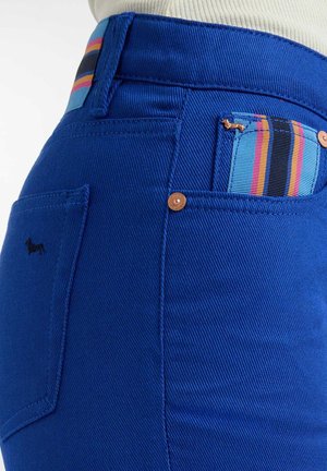 Jean en denim bleu avec une coupe ajustée, des coutures visibles et un accent distinctif en tissu rayé sur la poche de la ceinture.