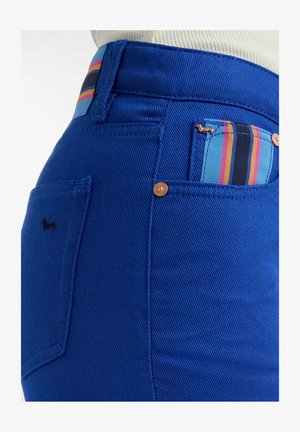 Los jeans de mezclilla azul presentan un diseño ajustado, costuras visibles y un distintivo detalle de tela a rayas en el bolsillo de la cintura.