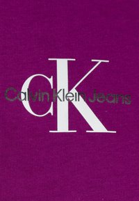 Fialová látka s textem "Calvin Klein Jeans" v šedé barvě a výrazným bílým logem "CK". Jemná textura.