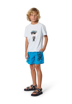 Băiat blond purtând un tricou alb cu imprimeu grafic, pantaloni scurți de un albastru aprins cu textul "Karl Lagerfeld" și sandale negre cu bretele inscripționate cu numele mărcii.