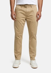 Chinos beige à texture lisse, affichant une coupe droite, deux plis à l'avant, et une boucle pour ceinture, associés à des baskets blanches.