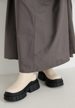 Les bottines à plateforme beiges présentent une semelle noire épaisse, une tige en cuir lisse et un côté élastique. Elles sont associées à une jupe grise.