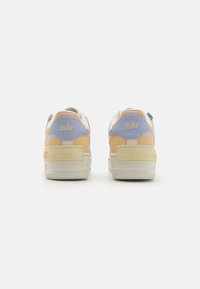 Zapatillas Nike con una base blanca, acentos en naranja pastel, amarillo y azul claro. Presentan una suela texturizada y una marca en el talón.