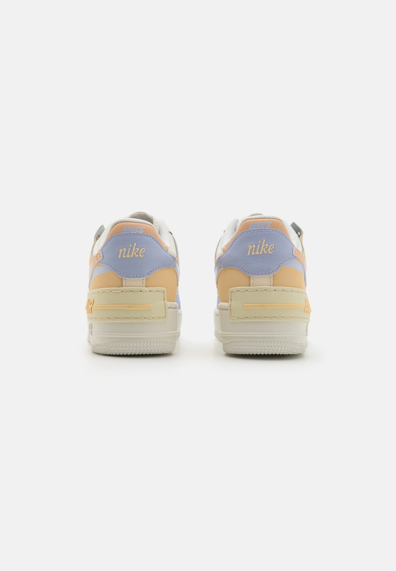Zapatillas Nike con una base blanca, acentos en naranja pastel, amarillo y azul claro. Presentan una suela texturizada y una marca en el talón.