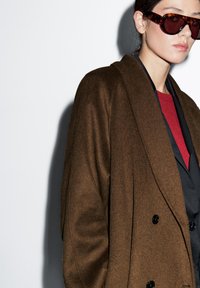 Brązowy wełniany płaszcz o podwójnym zapięciu, w połączeniu z czarną warstwą wewnętrzną i czerwoną górą. Model nosi ciemne, oversize'owe okulary przeciwsłoneczne.