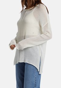 Leichter, gerippter Strickpullover in Creme mit langen Ärmeln, seitlichen Schlitzen und lockerer Passform. Strukturierter Stoff mit leicht durchscheinendem Effekt.