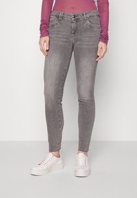 Persoon in versleten grijze skinny jeans, een lange paarse top en witte platform sneakers, staand tegen een effen achtergrond.