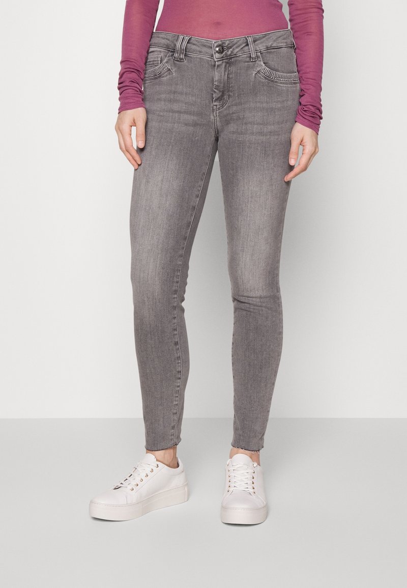 Persoon in versleten grijze skinny jeans, een lange paarse top en witte platform sneakers, staand tegen een effen achtergrond.